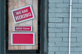 job recruitment We Are Hiring Sign in Window of Brick Building - A red and white sign in a window of a brick building reads We Are Hiring with a smaller sign that says Apply Today.