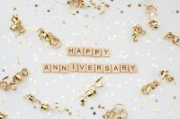 celebration anniversary The words 'HAPPY ANNIVERSARY' in wooden letter tiles, surrounded by gold and silver coloured stars and gold curls, all set against a white background. 