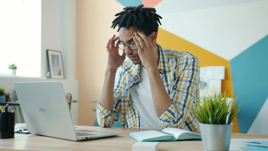 Chronic fatigue Exhausted mixed race man is feeling headache working with laptop in office room touching head suffering from migraine. People and health concept. 