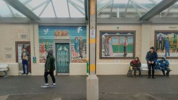 mosaic community Bray Station A colourful and charming scene captured at the railway station in Bray, Co. Wicklow. The image is framed by two large, vibrant mosaics depicting local, historical figures and scenes, related to the station's heritage. A pedestrian passes by in motion blur on the left, while a group of people watch on, waiting on the right, all under the sheltered platform roof.