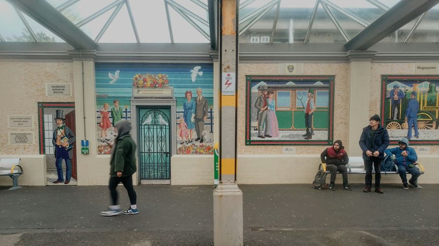 mosaic community Bray Station A colourful and charming scene captured at the railway station in Bray, Co. Wicklow. The image is framed by two large, vibrant mosaics depicting local, historical figures and scenes, related to the station's heritage. A pedestrian passes by in motion blur on the left, while a group of people watch on, waiting on the right, all under the sheltered platform roof. 