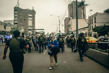 City conflict A line of police officers in riot gear marches down the street, forming a barrier between themselves and protesters. A photographer walks ahead of them, capturing the unfolding events.