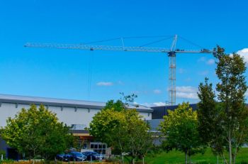 Construction industry This photograph captures a construction crane towering over a modern building, set against a clear blue sky and surrounded by lush green trees. The image highlights the intersection between urban development and natural elements, making it ideal for content related to construction, architecture, urban planning, or environmental balance. The vibrant colors and clean composition provide a dynamic view of city growth and development. canada,bc,langley