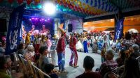 Party Theater Costumed reisado performers in elaborate headdresses interact with the audience on the floor of the Polo Repente venue during São João de Caruaru, as spectators seated in chairs watch under a canopy of multicolored paper flags and a large illustrated stage backdrop. 