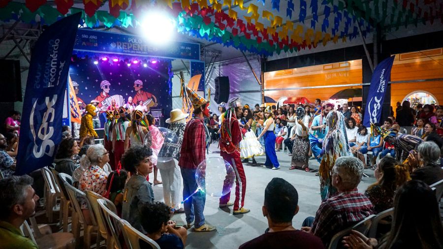 Party Theater Costumed reisado performers in elaborate headdresses interact with the audience on the floor of the Polo Repente venue during São João de Caruaru, as spectators seated in chairs watch under a canopy of multicolored paper flags and a large illustrated stage backdrop. 