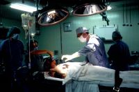 Criminal Operation Biopsy. A Caucasian woman patient is being prepared for surgery. Her left breast is visible and anesthesia is being administered. The male physician has his hand extended and is examining the patient. Several operative attendants are also visible. A surgical biopsy is being performed to determine exact nature of solid tumor. Creator: Linda Bartlett surgery,health,cancer