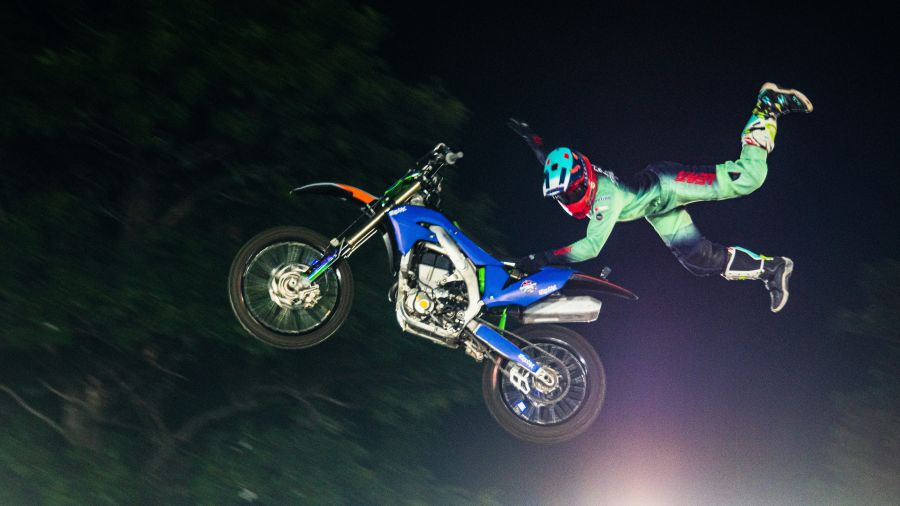 gravity performance A freestyle motocross rider performs an airborne trick at night, extending their body off the side of a blue dirt bike. The bike and rider are suspended high in the air against a dark background, emphasizing motion and athletic skill. 