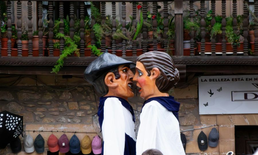 Giants Festival Two large paper-mâché figures, known as "Cabezudos," representing a man with a black beret and a woman with brown hair, stand facing each other in a profile view. They are dressed in white shirts with blue scarves. The figures are positioned in front of a stone building featuring a dark wooden balcony filled with various potted plants. Below the balcony, several baseball caps are displayed for sale against the stone wall. 