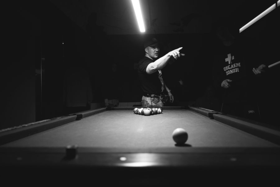 tension sports A cinematic black-and-white image of a pool table moments before the break — an overhead light isolates the racked balls and cue ball while a focused player points and directs, creating tense, low-key atmosphere in the dark pool room. 
