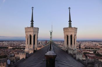Lyon debate Symmetrical dusk view from the roof of Lyon’s Basilica of Notre-Dame de Fourvière: the twin campanile towers and the archangel statue align over the city, with pastel sunset tones fading across the skyline. A serene, elevated perspective blending sacred architecture and sweeping urban panorama. 