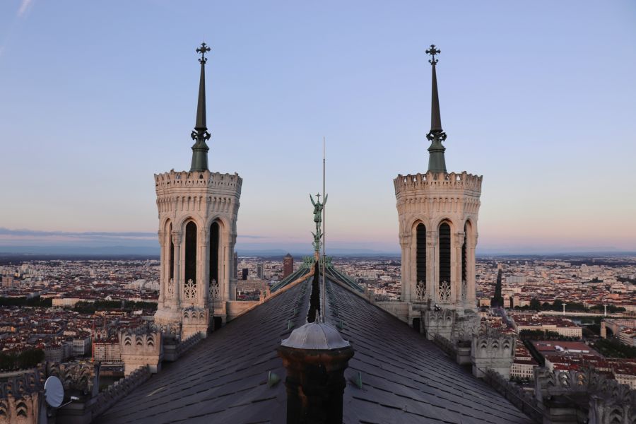 Lyon debate Symmetrical dusk view from the roof of Lyon’s Basilica of Notre-Dame de Fourvière: the twin campanile towers and the archangel statue align over the city, with pastel sunset tones fading across the skyline. A serene, elevated perspective blending sacred architecture and sweeping urban panorama. 