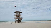 Coastal surveillance This photograph captures a wooden lifeguard tower standing tall on a sandy beach. A red flag, indicating caution due to hazardous conditions, is flying atop the tower. The sky above is partly cloudy with a pattern of altocumulus clouds, and the ocean in the background appears choppy. The beach is nearly empty, adding to the sense of caution suggested by the red flag. grey,sandy beach,caution at beach