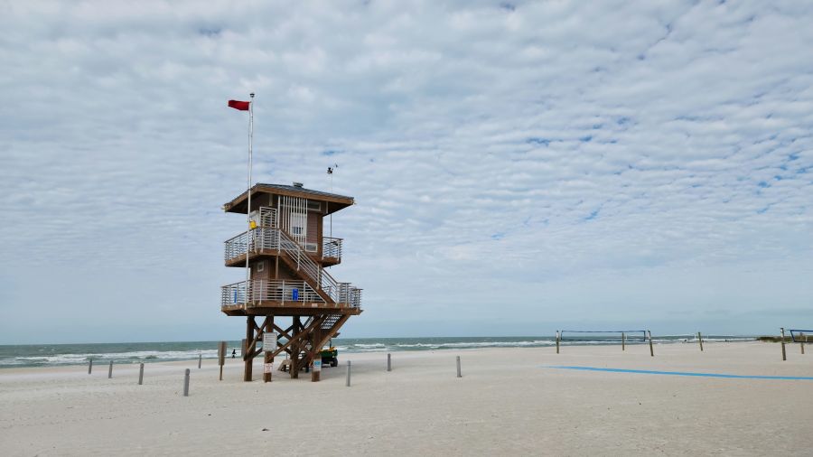 Coastal surveillance This photograph captures a wooden lifeguard tower standing tall on a sandy beach. A red flag, indicating caution due to hazardous conditions, is flying atop the tower. The sky above is partly cloudy with a pattern of altocumulus clouds, and the ocean in the background appears choppy. The beach is nearly empty, adding to the sense of caution suggested by the red flag. grey,sandy beach,caution at beach