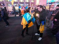 war journalist A woman draped in a signed blue and yellow Ukrainian flag gives an interview on a busy Times Square sidewalk at night. A man holds a small light and phone on a tripod to film her while demonstrators and onlookers gather around. Protesters rally in Times Square, New York City, on February 15, 2022, standing in solidarity with Ukraine and speaking out against the war at an anti-war demonstration. Photo: Olek Buzunov (@buzunov.photoart) 