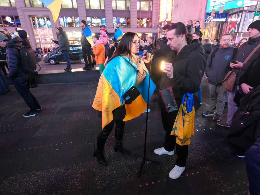 war journalist A woman draped in a signed blue and yellow Ukrainian flag gives an interview on a busy Times Square sidewalk at night. A man holds a small light and phone on a tripod to film her while demonstrators and onlookers gather around. Protesters rally in Times Square, New York City, on February 15, 2022, standing in solidarity with Ukraine and speaking out against the war at an anti-war demonstration. Photo: Olek Buzunov (@buzunov.photoart) 