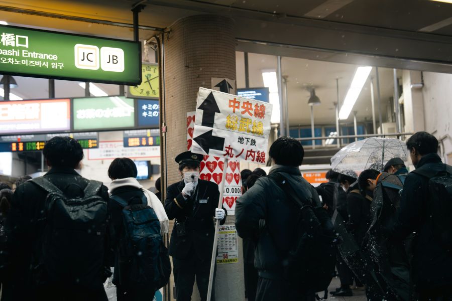 Train delays In the midst of a heavy snowfall at Ochanomizu Station, Tokyo, a station attendant diligently informs passengers of train schedule disruptions while guiding them to alternative transportation options. urban commute,rail network,schedule changes