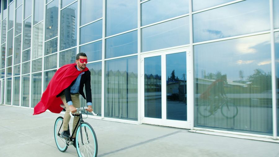 Heroic action Slow motion of superhero businessman riding bike in business district in city street wearing red cape and mask. Eco transport and work achievements concept. 