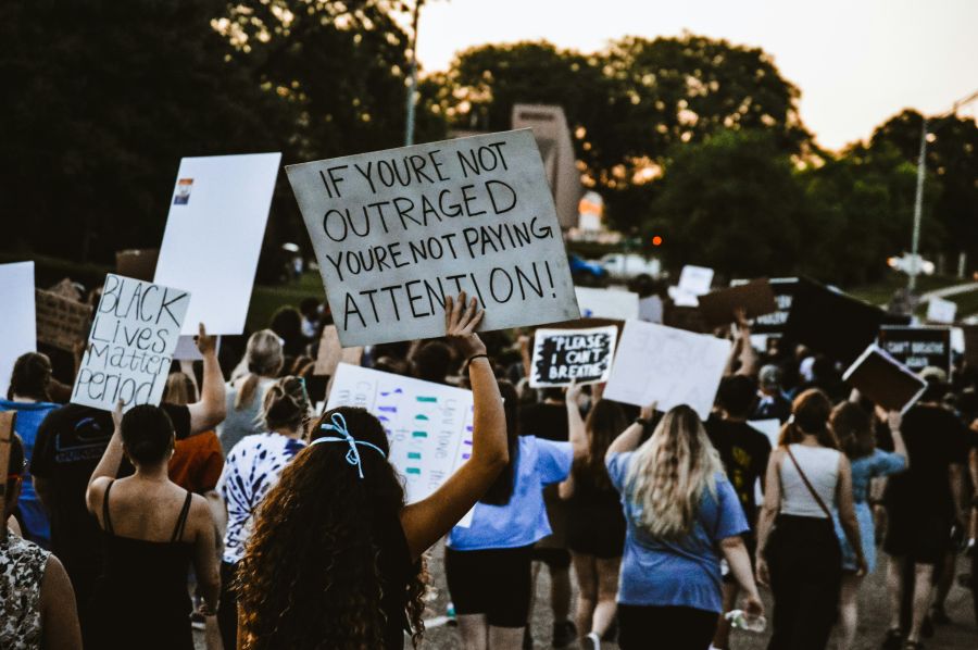 discrimination protest scene from the 2020 summer protests in the streets of Nebraska, with a girl holding a sign that says "if you're not outraged, you're not paying attention"  