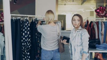 thief crime Young woman stealing wallet from female customer choosing clothes in shop. Thief looking around carefully and walking away. Fine boutique with clothing, bags and shoes in background 