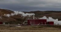 Geothermal energy An industrial geothermal power plant set in a barren landscape, with steam plumes emitted from pipes and silos against a backdrop of hilly terrain. 