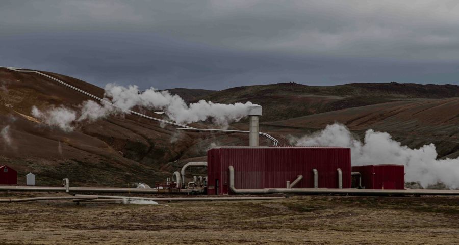 Geothermal energy An industrial geothermal power plant set in a barren landscape, with steam plumes emitted from pipes and silos against a backdrop of hilly terrain. 