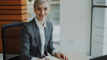 Woman candidate Portrait of Attractive blonde businesswoman sitting at table writing in notebook smiling into camera in modern office indoors