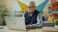 retirement work Portrait of handsome old man in glasses using laptop typing at home then looking at camera and smiling. Modern technology and retirement concept. 
