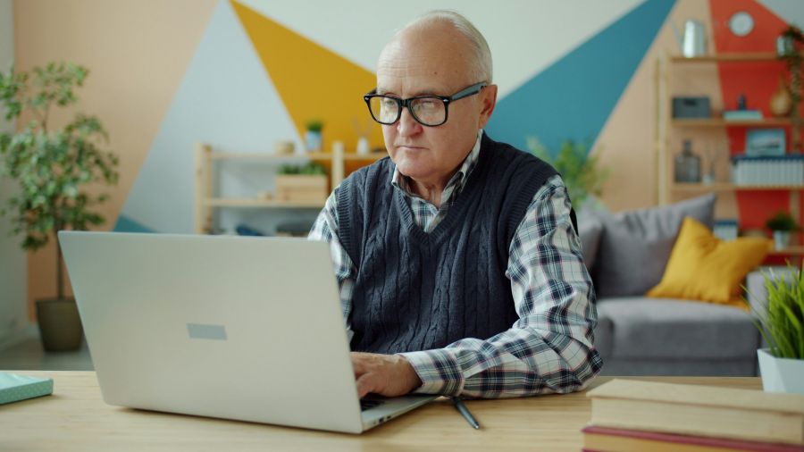 retirement work Portrait of handsome old man in glasses using laptop typing at home then looking at camera and smiling. Modern technology and retirement concept. 