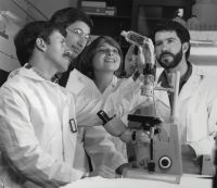 scientific discovery A group of scientists, 1 woman and 3 men, gather around a microscope to examine a tissue culture flask. 