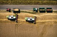 Farm vehicles Aerial views of two combine harvesters operating in a wheat field, transferring harvested grain into trailers pulled by tractors parked along an adjacent rural road. 