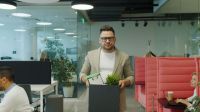 Job loss Fired middle-aged man in suit is walking in shared office leaving workplace with box of belongings feelings sad being unemployed. Dismissal and emotions concept. 
