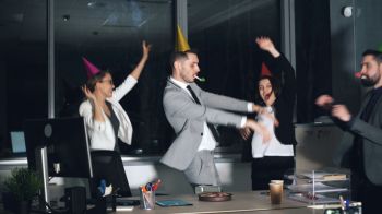 celebration interview Happy young people office workers in suits are dancing at company party wearing party hats and blowing horns. Birthday cake and gift box are visible on desk.