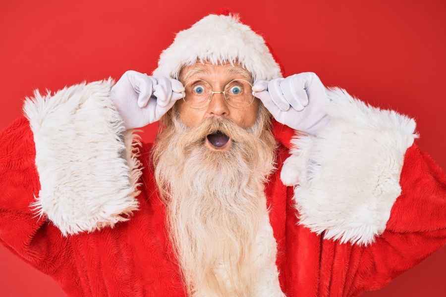 Senior workforce Old senior man with grey hair and long beard wearing santa claus costume holding glasses afraid and shocked with surprise and amazed expression, fear and excited face.  senior,emotional,expressive