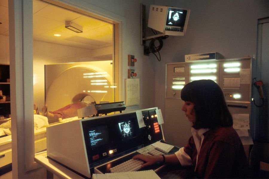 Radiology healthcare A computer-assisted tomographic (CAT) scanner, with a Caucasian female technician working at a screen and behind a glass wall. A patient is on a table and being tested by the CAT scanner. The lighting is very subdued. This new technology revolutionized detection of brain tumors. 