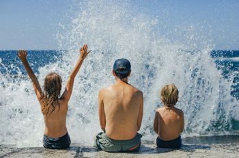 vacation kids Two children and one adult man get splashed by the ocean spray, one child holds their hands in the air to embrace the joy of the wave
