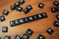 feminism violence Black and white Scrabble wood letter blocks form the word FEMINIST on a wooden table top surrounded by other game pieces. 