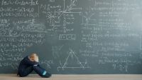 Exam stress Sad child in suit is sitting on floor near black chalkboard wall with formulas thinking solving problem alone. Education, childhood and emotions concept. 
