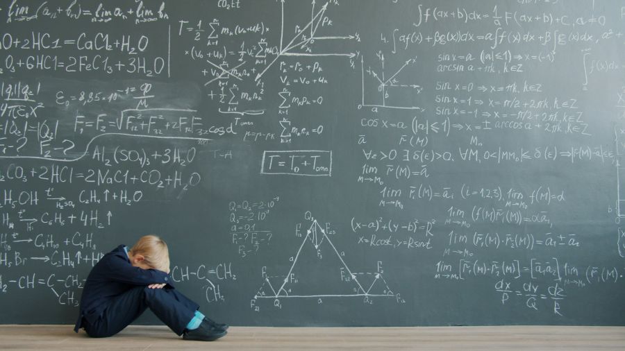 Exam stress Sad child in suit is sitting on floor near black chalkboard wall with formulas thinking solving problem alone. Education, childhood and emotions concept. 